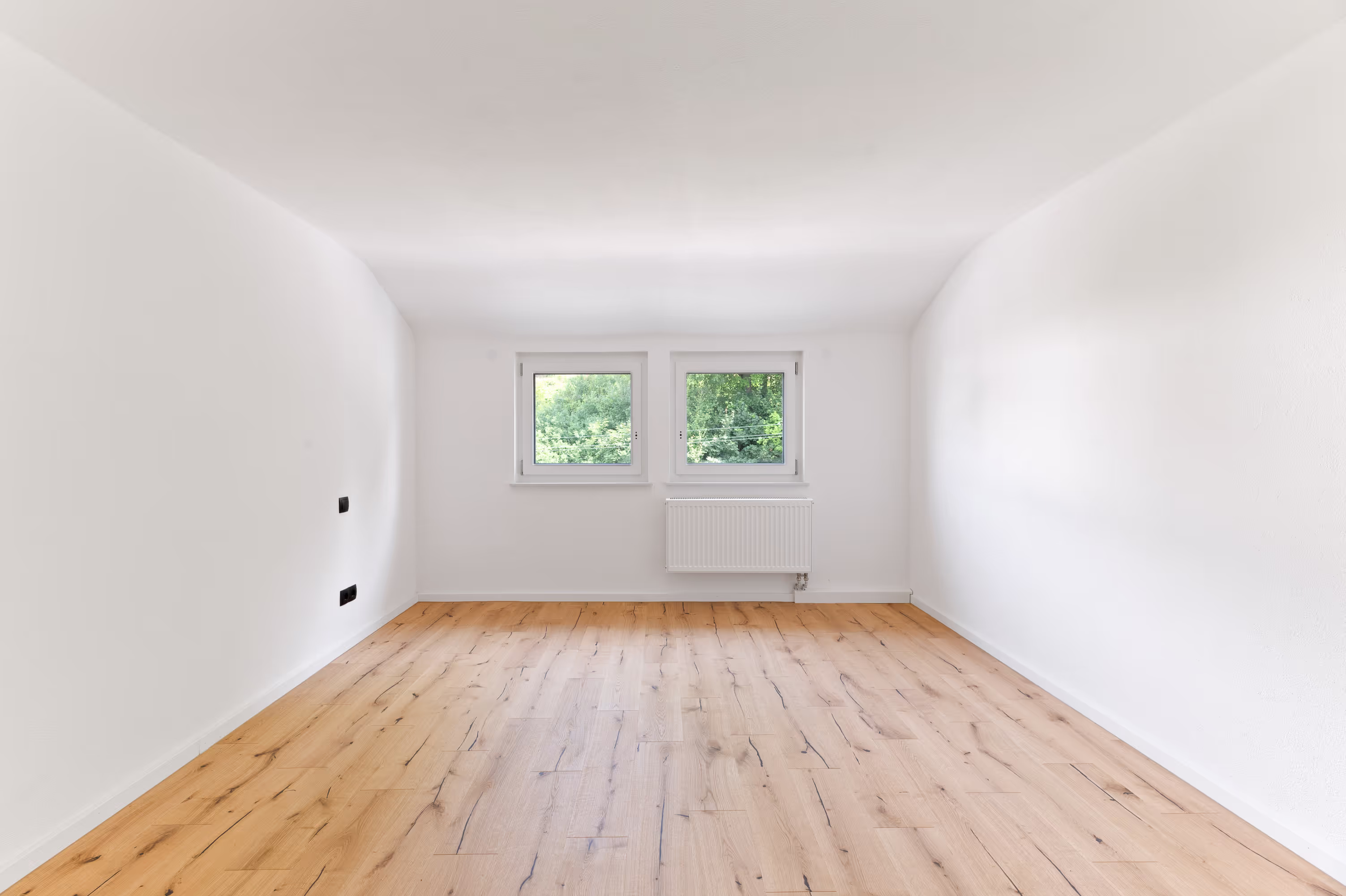 Bright, empty room with two windows, wooden flooring, and a white radiator beneath the windows, offering a view of greenery outside.