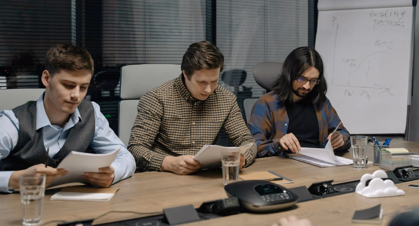 Three young professionals reviewing documents together at a conference table during a focused planning meeting, with a whiteboard visible in the background showing project notes