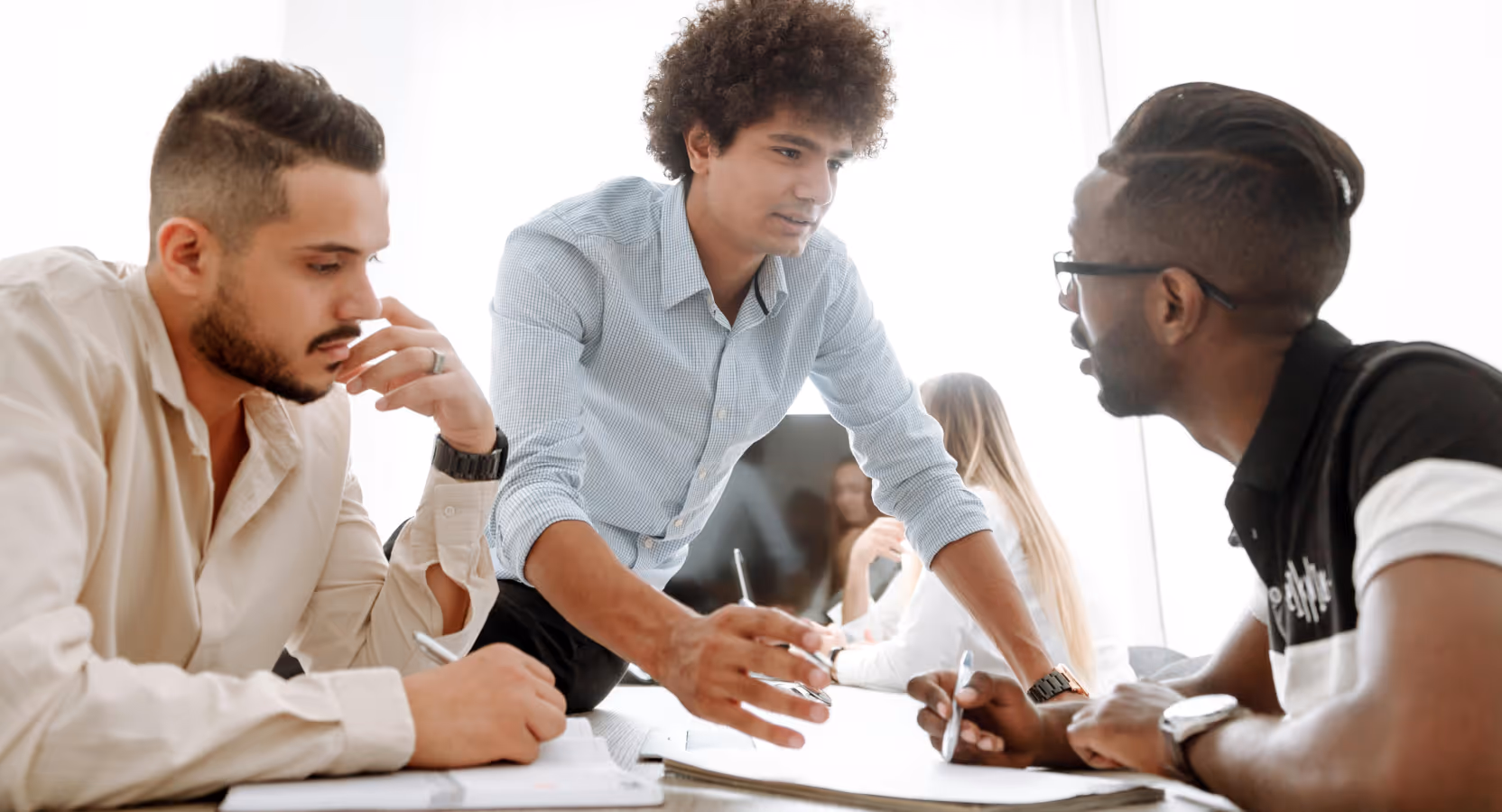 A small team in a bright office environment working together on documents, with a person in a blue checkered shirt writing on paper while others observe and participate in the meeting