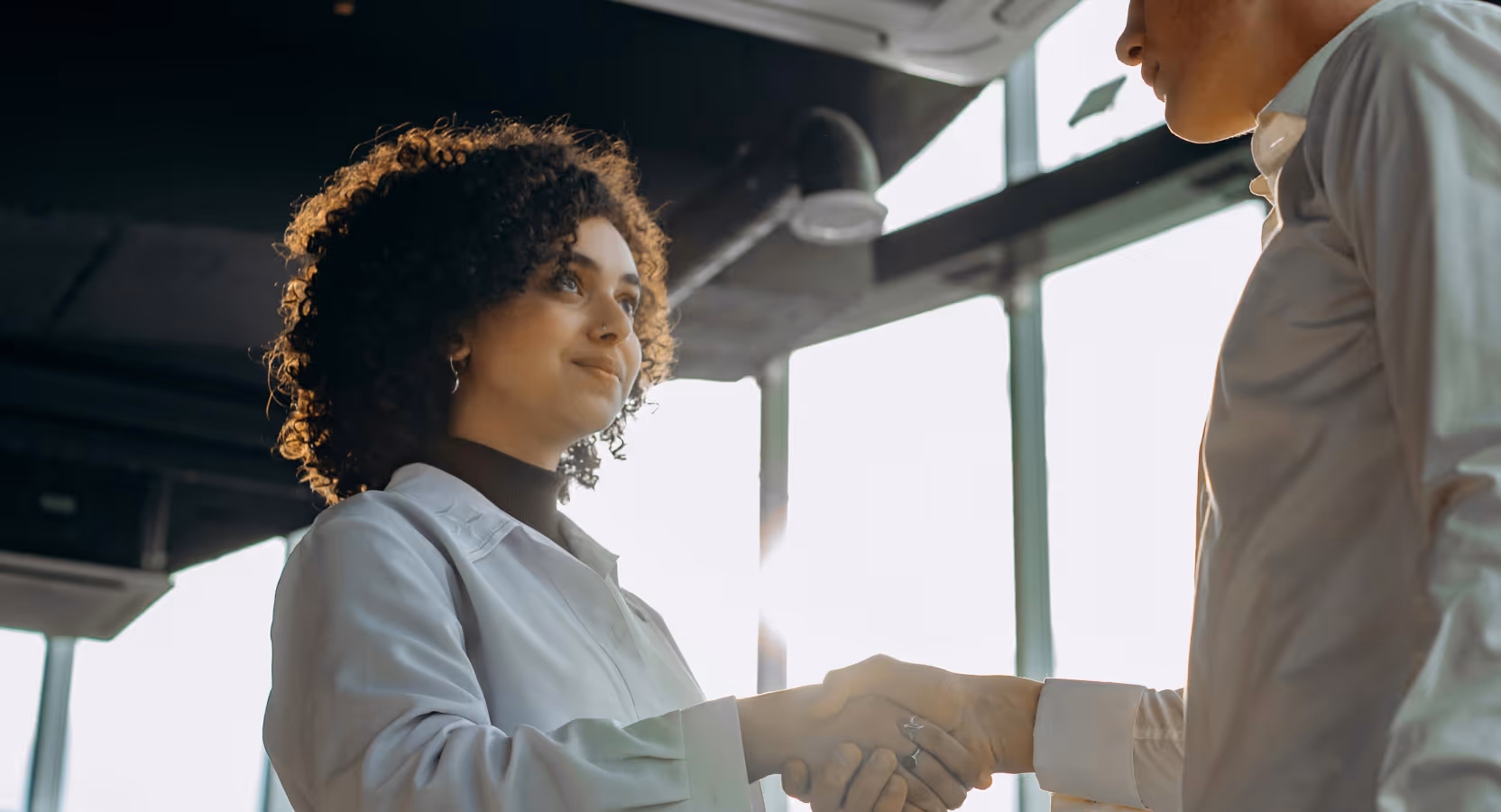 Two business professionals in white shirts shaking hands in a modern office with large windows, representing partnership in HealthTech development