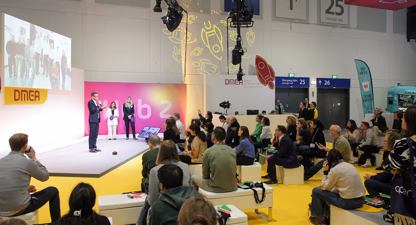 Startup pitch session at the DMEA Berlin 2025 innovation hub, with a lively audience and presenters on a yellow-themed stage