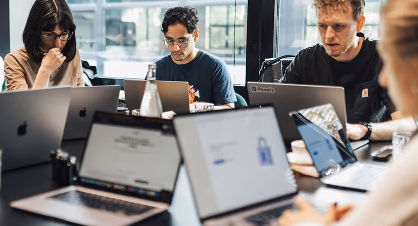 Group of people working together at a shared table, each focused on their laptops, suggesting collaboration on a technology or software project.