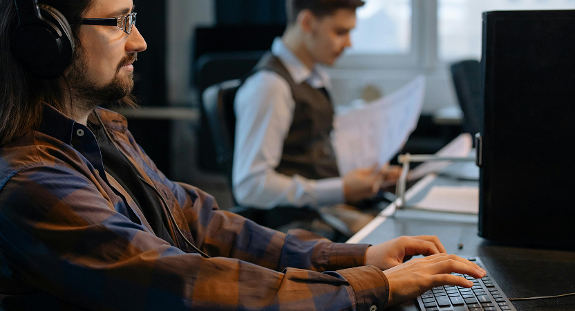 Developer wearing headphones types at a desktop while a teammate reviews documents in the background—real-world use of AI coding tools.
