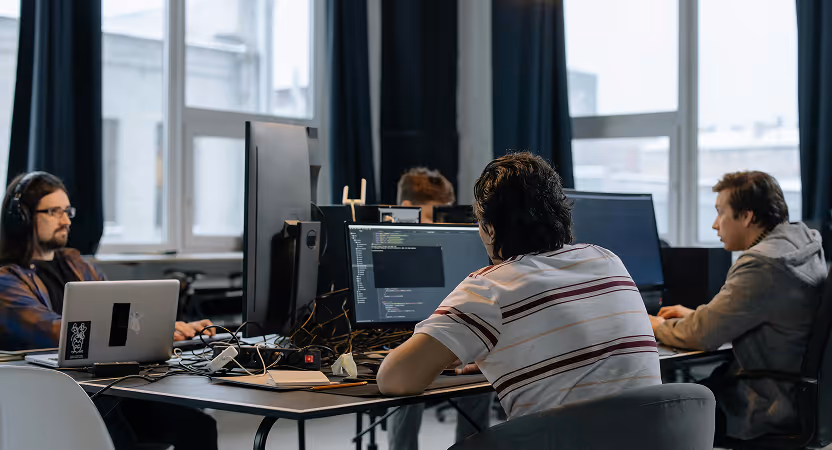 Software team coding at shared desks with multiple monitors, collaborating on production workflows using AI coding assistants.