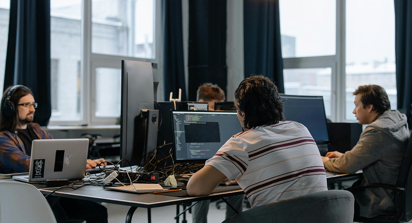Software team coding at shared desks with multiple monitors, collaborating on production workflows using AI coding assistants.