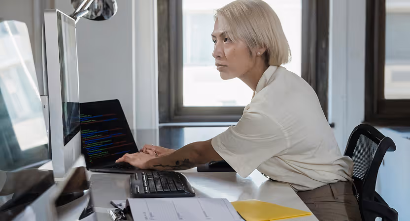Engineer reviews code on a laptop and external monitor in a modern office, validating AI-generated changes before deployment.
