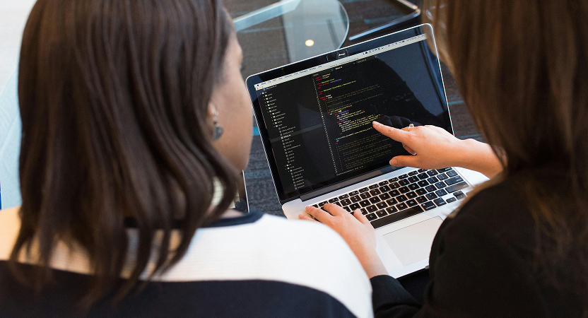 Two female developers collaborating on healthcare AI code, with one pointing at laptop screen during technical review