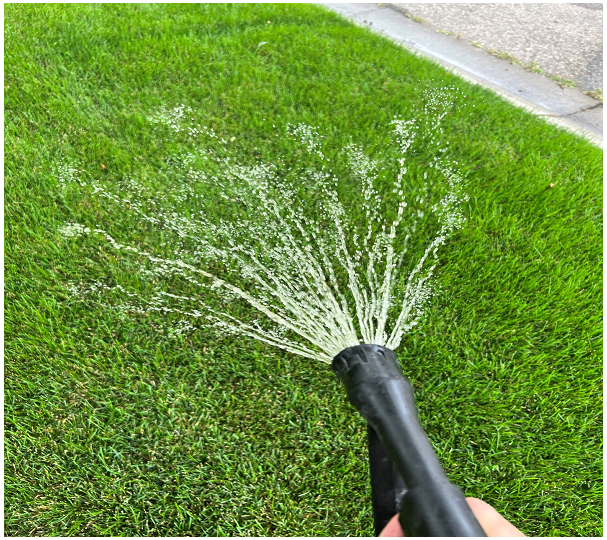Hand holding a garden hose spraying water in multiple streams onto green grass near a sidewalk.