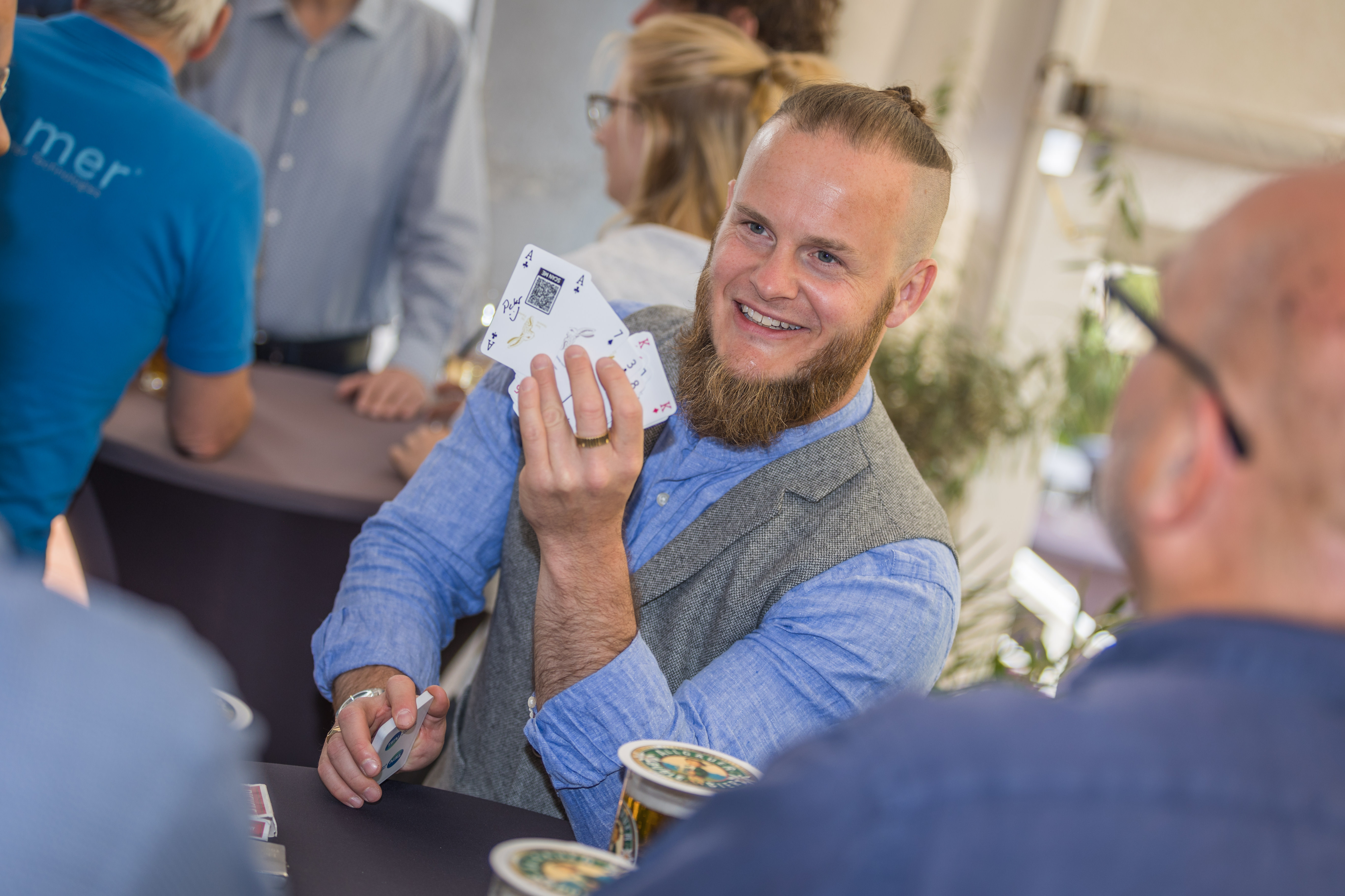 Ein lächelnder Mann mit Bart und hochgebundenem Haar hält Spielkarten in der Hand und sitzt an einem Tisch mit anderen Personen.