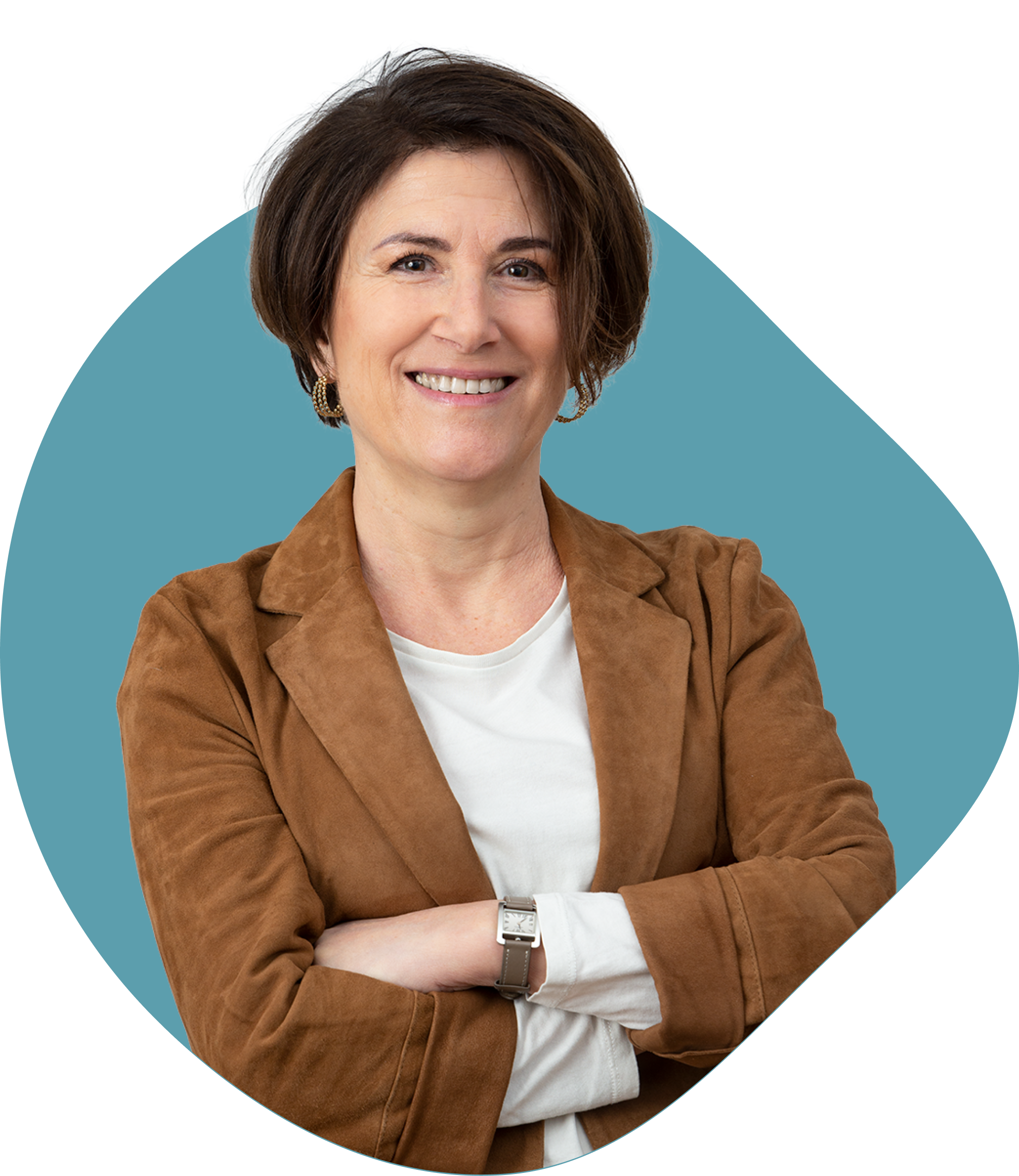 Femme souriante aux cheveux courts, veste marron et chemise blanche, bras croisés devant un fond bleu.