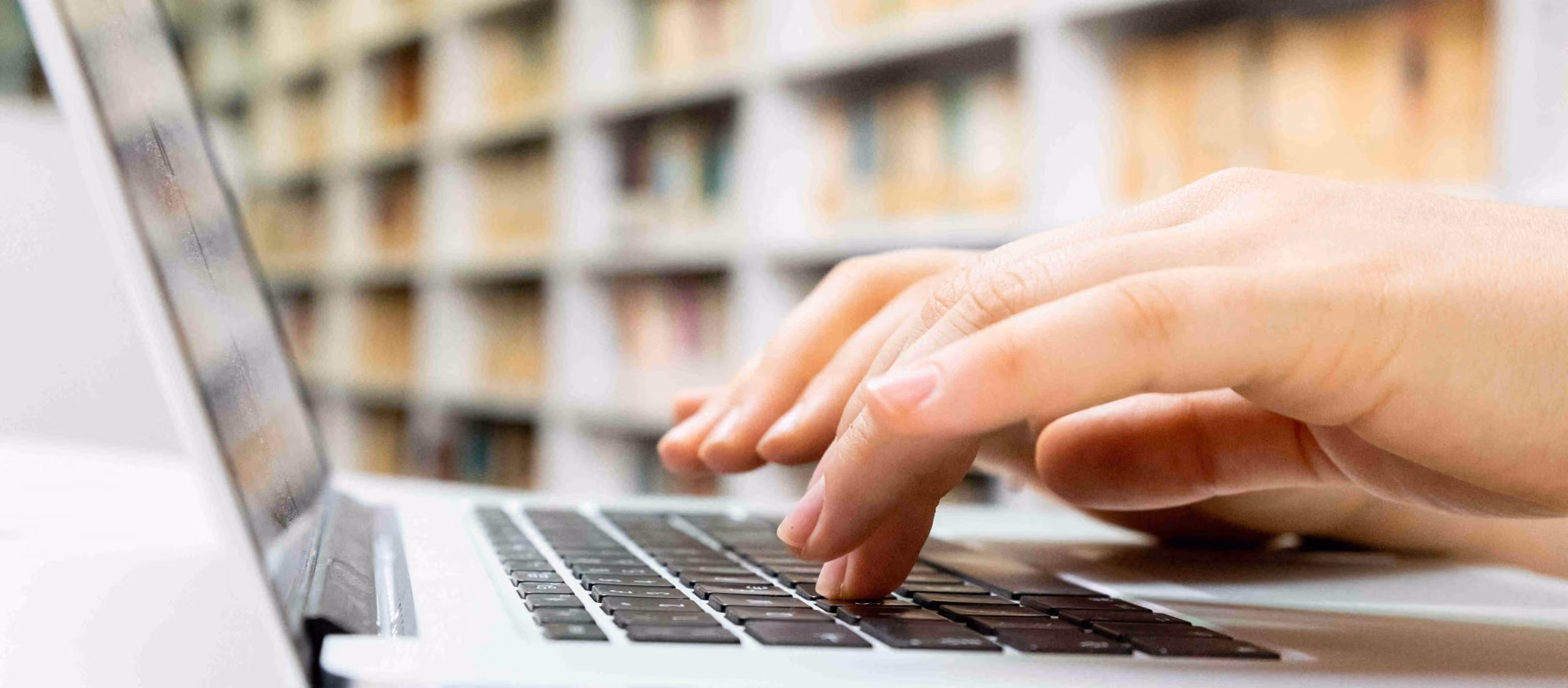 Manos escribiendo en un teclado de laptop con estantería de libros de fondo
