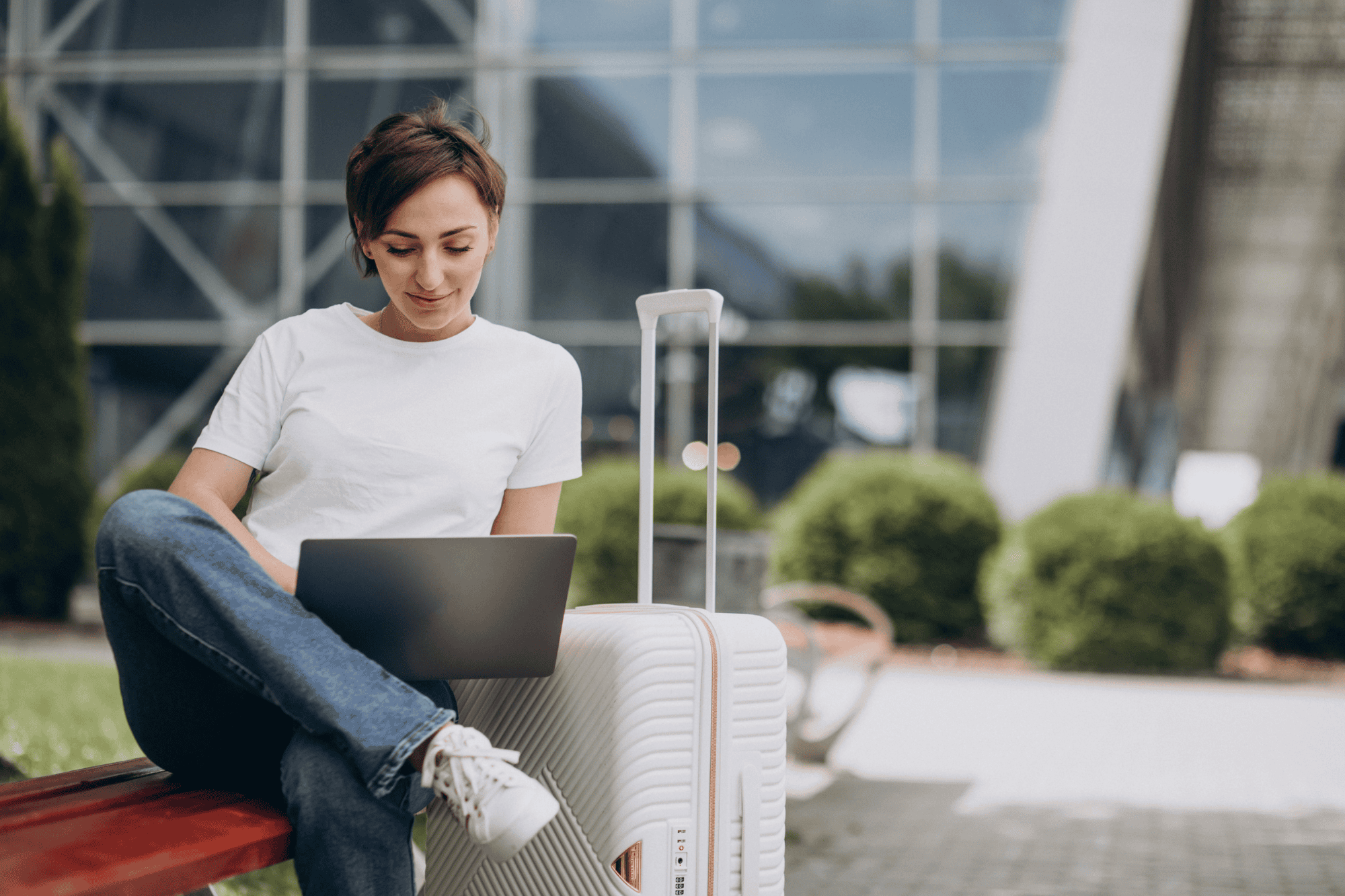 Woman working on laptop next to white suitcase outside building