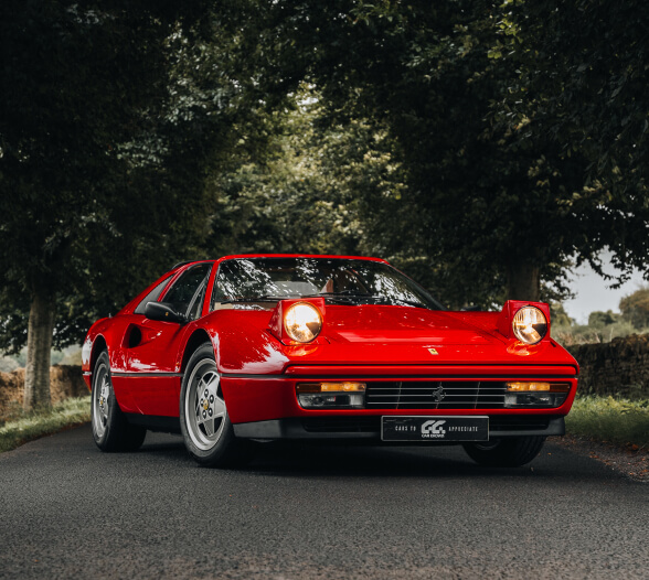 Red classic Ferrari sports car with pop-up headlights on a tree-lined road.