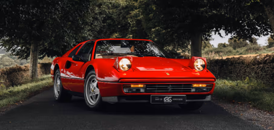 Red vintage sports car with pop-up headlights on a country road lined with trees and stone walls.