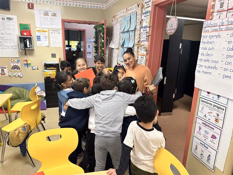 Students hugging their smiling teacher in a decorated elementary classroom