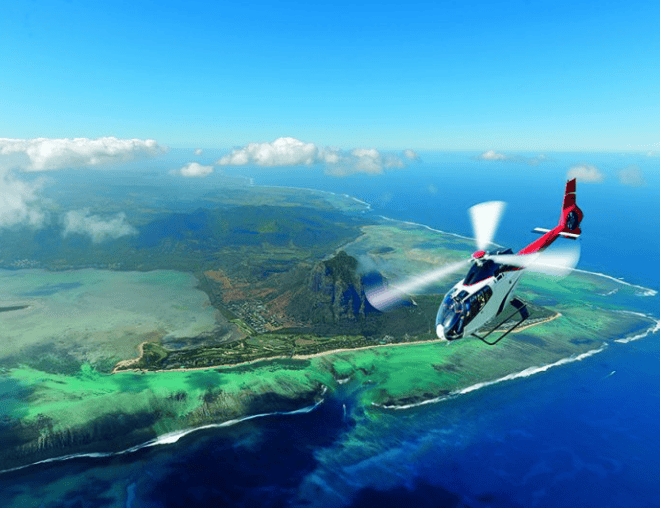 A white helicopter soaring above the emerald reefs and rugged volcanic landscape of Le Morne, Mauritius.