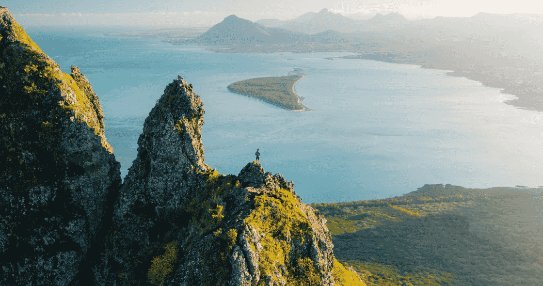Hiker standing on a rugged mountain peak overlooking a vast blue bay.