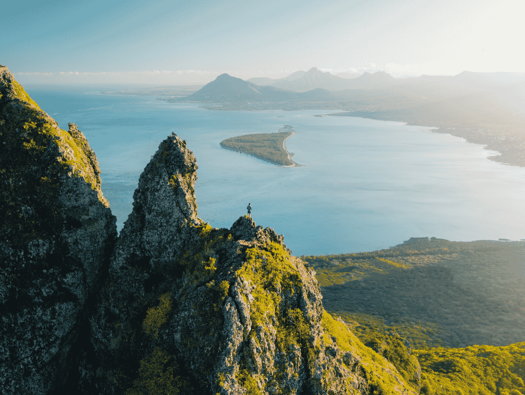 Hiker standing on a rugged mountain peak overlooking a vast blue bay.