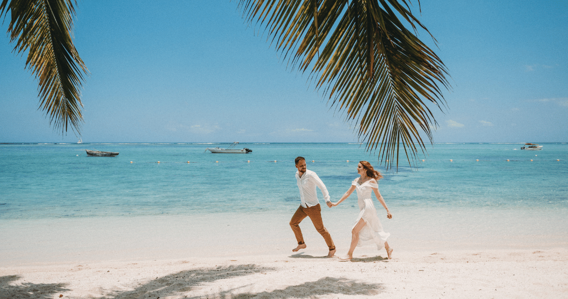 A man and woman running barefoot on a tropical beach, framed by palm tree leaves with boats in the ocean behind them.