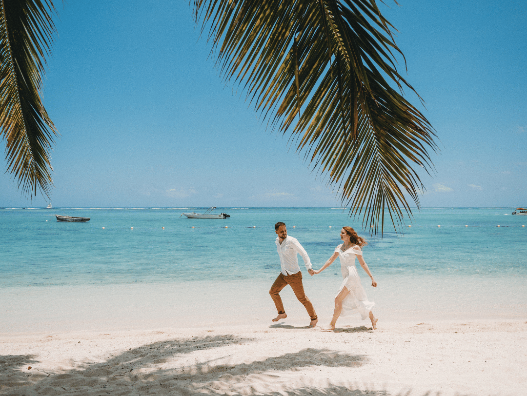 A man and woman running barefoot on a tropical beach, framed by palm tree leaves with boats in the ocean behind them.