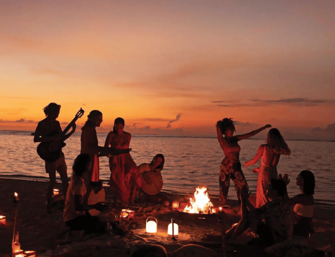 A sunset beach party featuring a warm bonfire, lit lanterns in the sand, and people socializing and playing instruments.