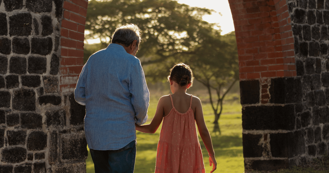 A young girl in a yellow dress walks joyfully alongside her grandfather.