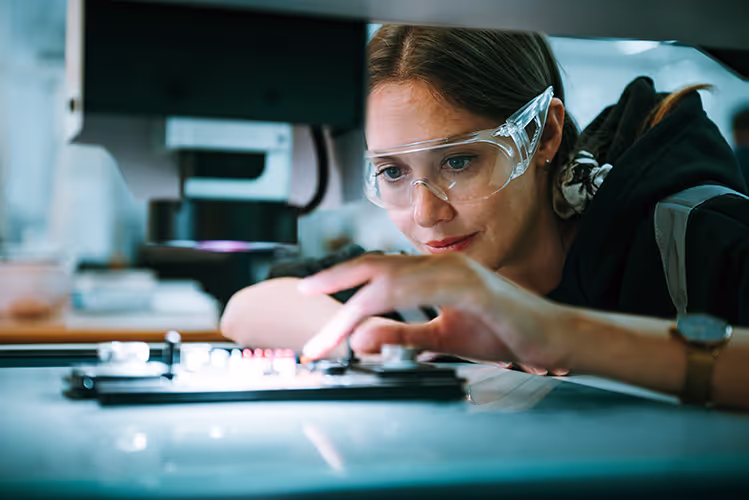 Frau mit Schutzbrille arbeitet konzentriert an einem Gerät in einem Labor.