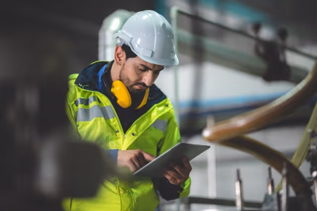Bautechniker mit weißem Schutzhelm und gelber Warnweste bedient ein Tablet in einer Industrieanlage.