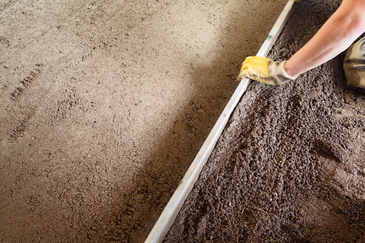 Nahaufnahme einer Person mit gelben Handschuhen, die glatten Estrich auf Bodenbelag mit einer Latte verteilt.