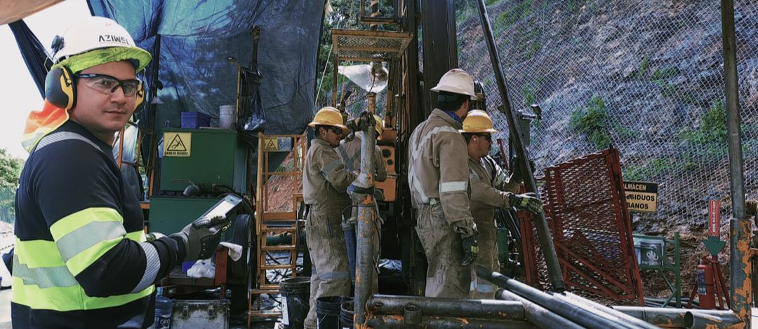 Un trabajador con una tableta en primer plano en un sitio de perforación, con tres compañeros operando una máquina ante una ladera con malla.