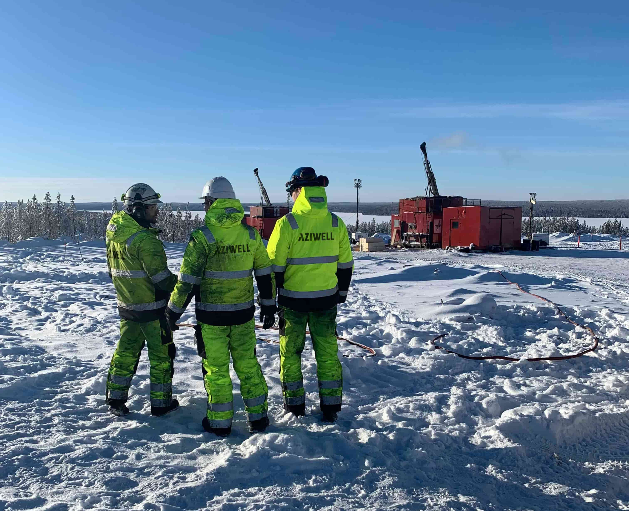 Three Aziwell workers standing at a snowy directional drilling site, looking towards two red drill rigs.