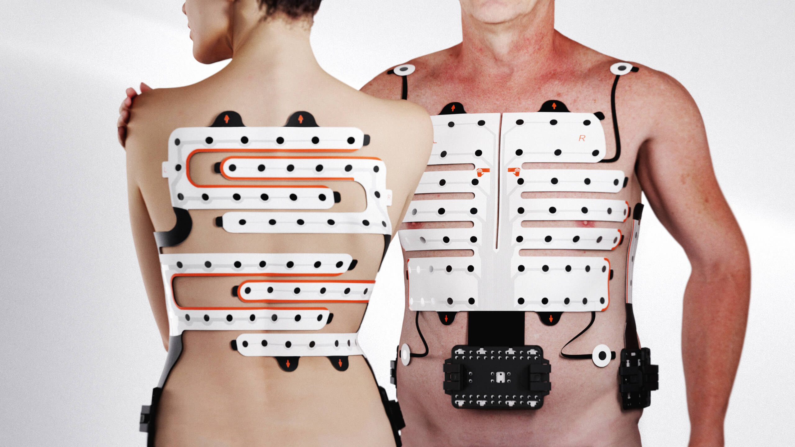 Two people, one facing forward and one backward, wearing white electronic sensor pads with black dots and orange lines attached to their torsos and backs, connected to black control units on the hips.