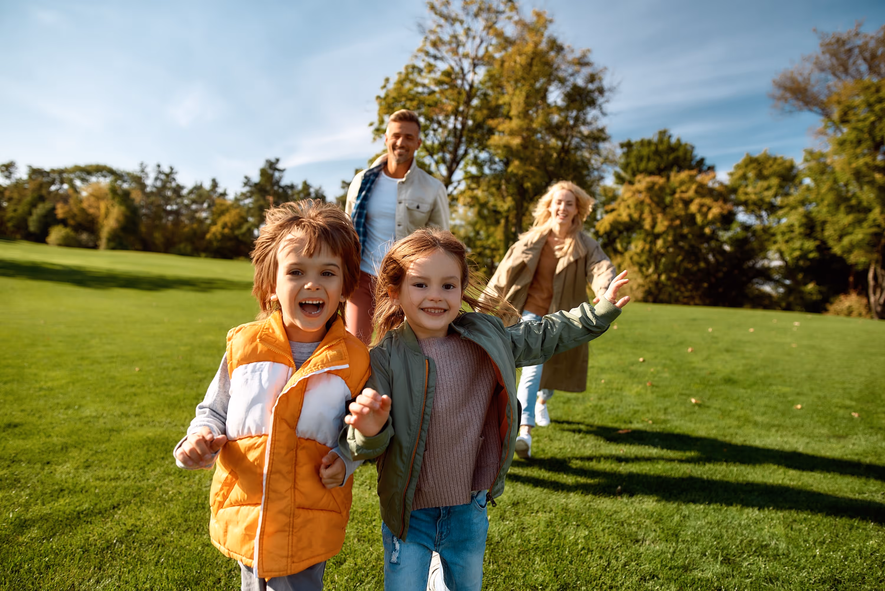 A happy family, with two young children running in the foreground, enjoying a sunny day in a park, representing the family-friendly lifestyle near The Abbey in Greenville, SC.