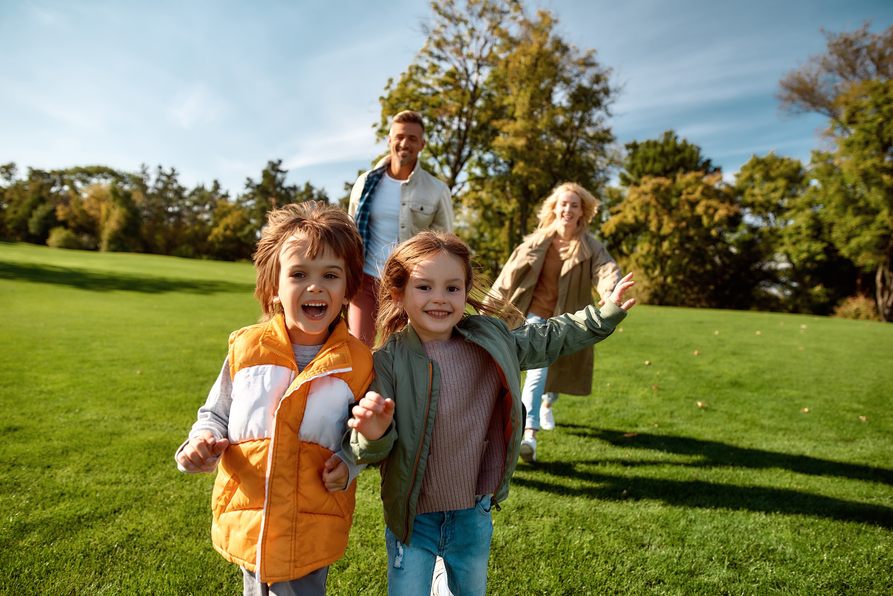A happy family, with two young children running in the foreground, enjoying a sunny day in a park, representing the family-friendly lifestyle near The Abbey in Greenville, SC.