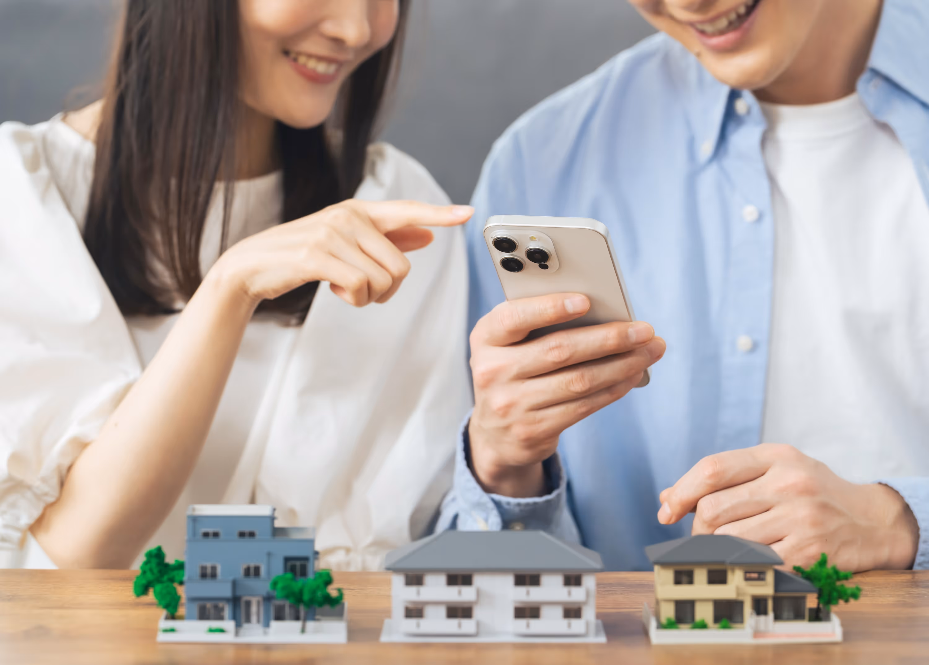 A couple smiling and pointing at a smartphone while looking at small house models, representing the convenient home search process for The Abbey in Greenville, SC.