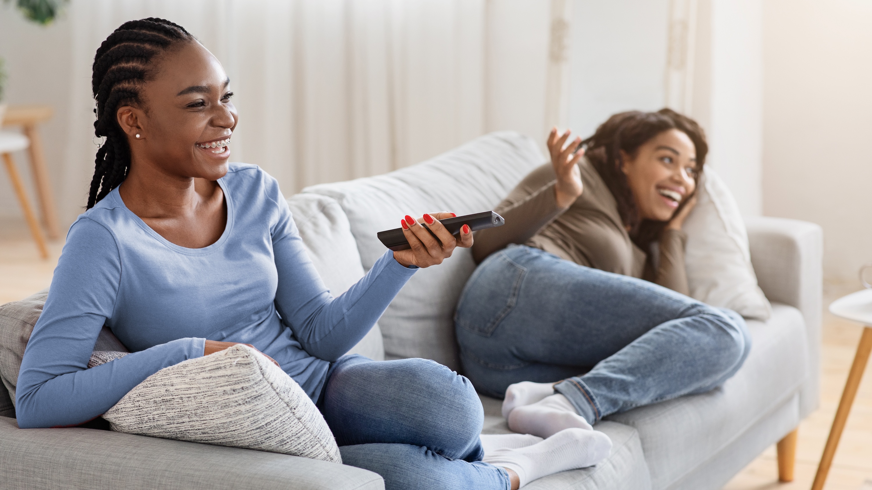 Two happy women laughing and watching TV on a comfortable sofa, representing the relaxing home life and entertainment amenities at The Abbey in Greenville, SC.