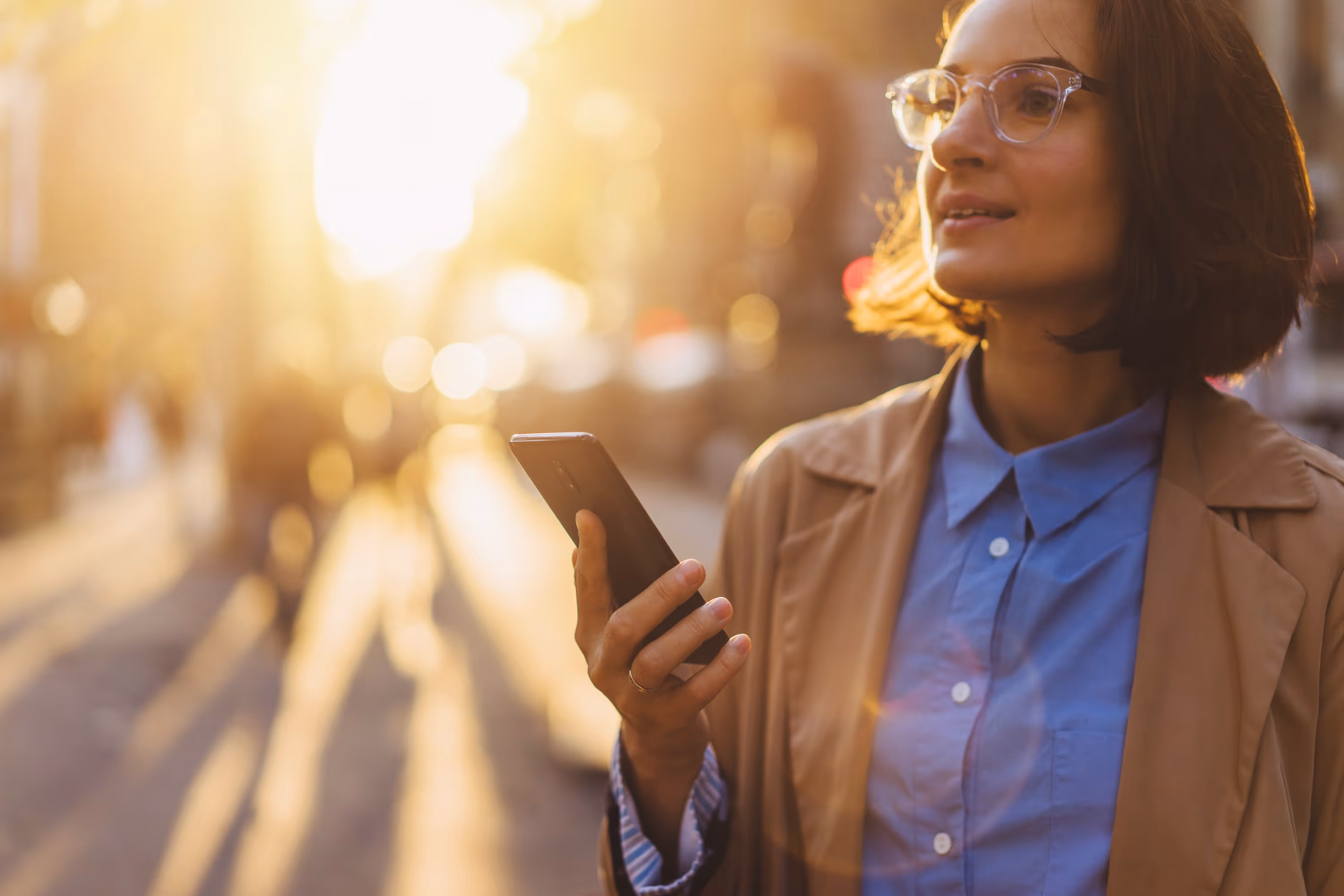 Professional woman in a trench coat holding a smartphone on a sunny city street in Greenville at sunset.