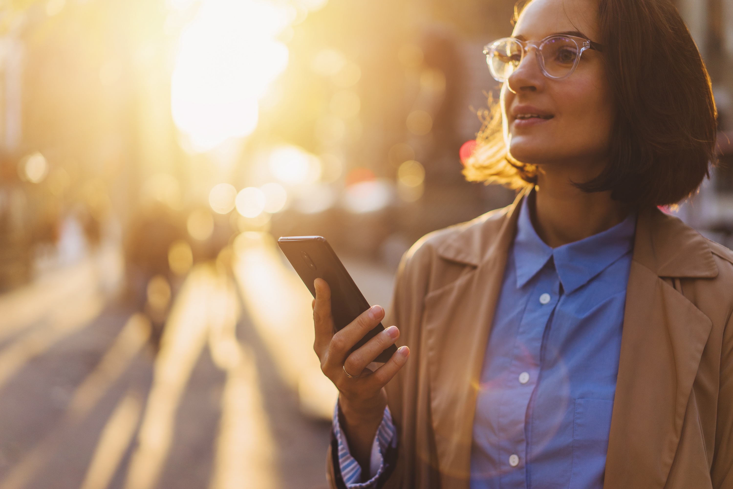 Professional woman in a trench coat holding a smartphone on a sunny city street in Greenville at sunset.