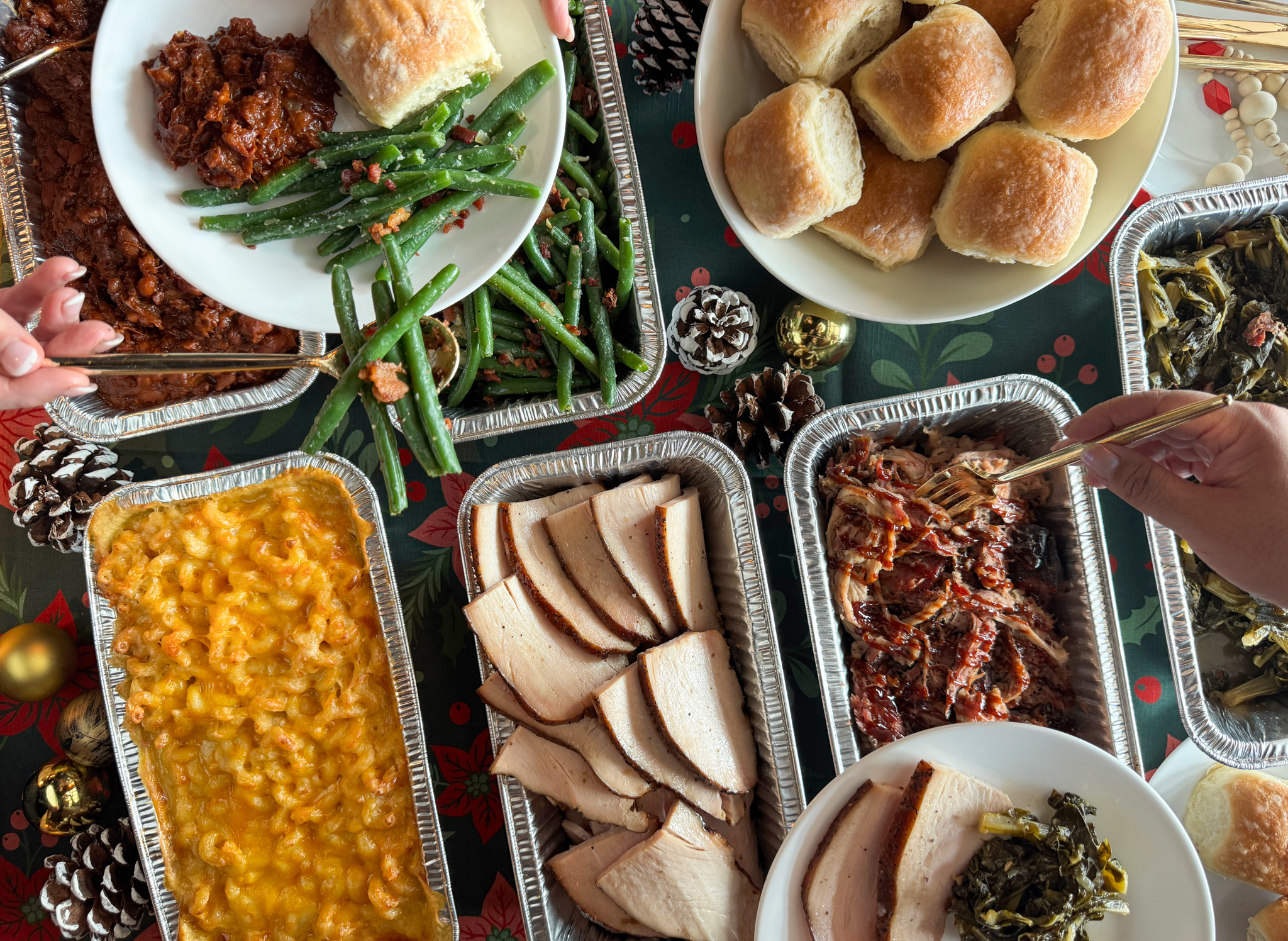 Christmas holiday meal spread with macaroni and cheese, green beans, baked beans, sliced turkey, pulled pork, collard greens, and dinner rolls on a festive tablecloth with pinecones and ornaments.