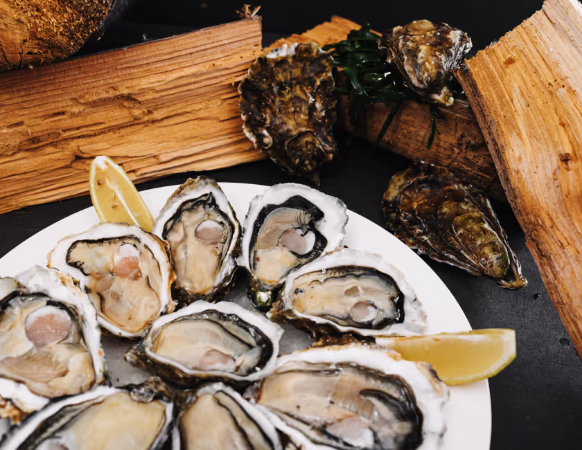 Een bord met verse open oesters en citroenpartjes, houtblokken en ongeopende oesters daarachter.