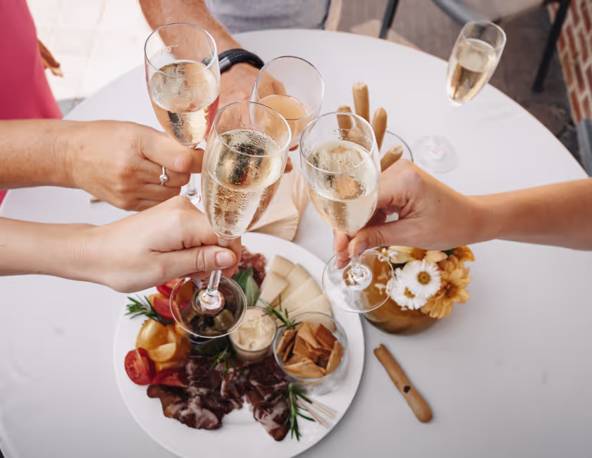 Mensen klinken met glazen champagne boven een tafel met een bord snacks en bloemen.