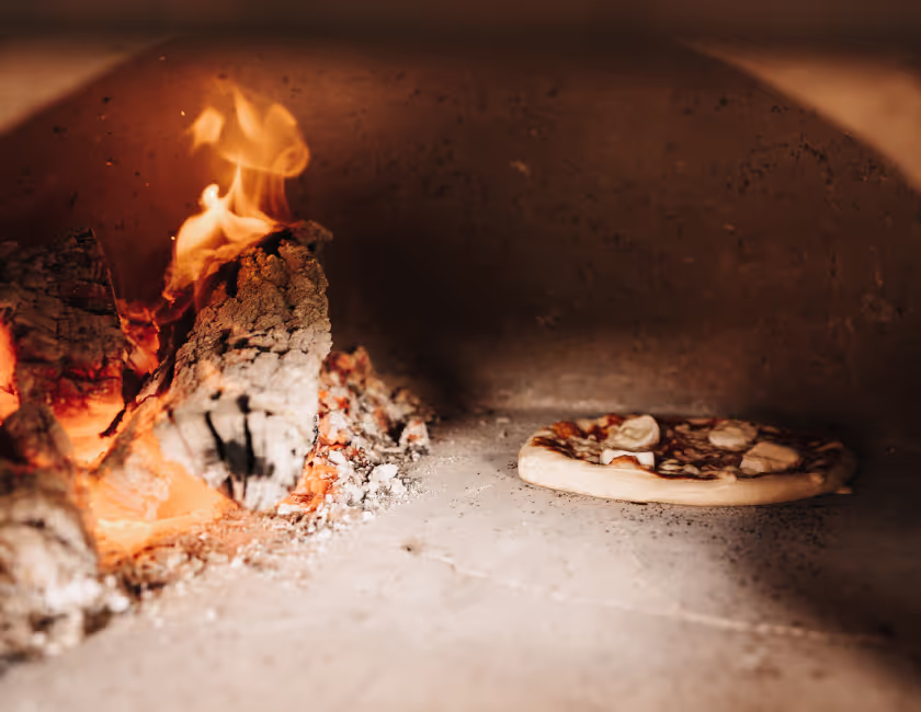 Een pizza bakt naast een vuur met brandend hout in een traditionele steenoven.