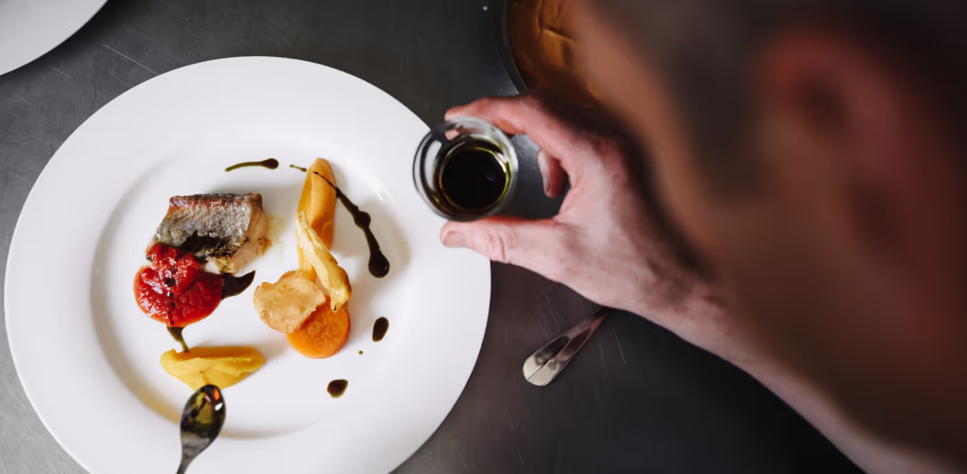Bord met een stuk vis en puree, naast hand die donkere saus op het gerecht giet.