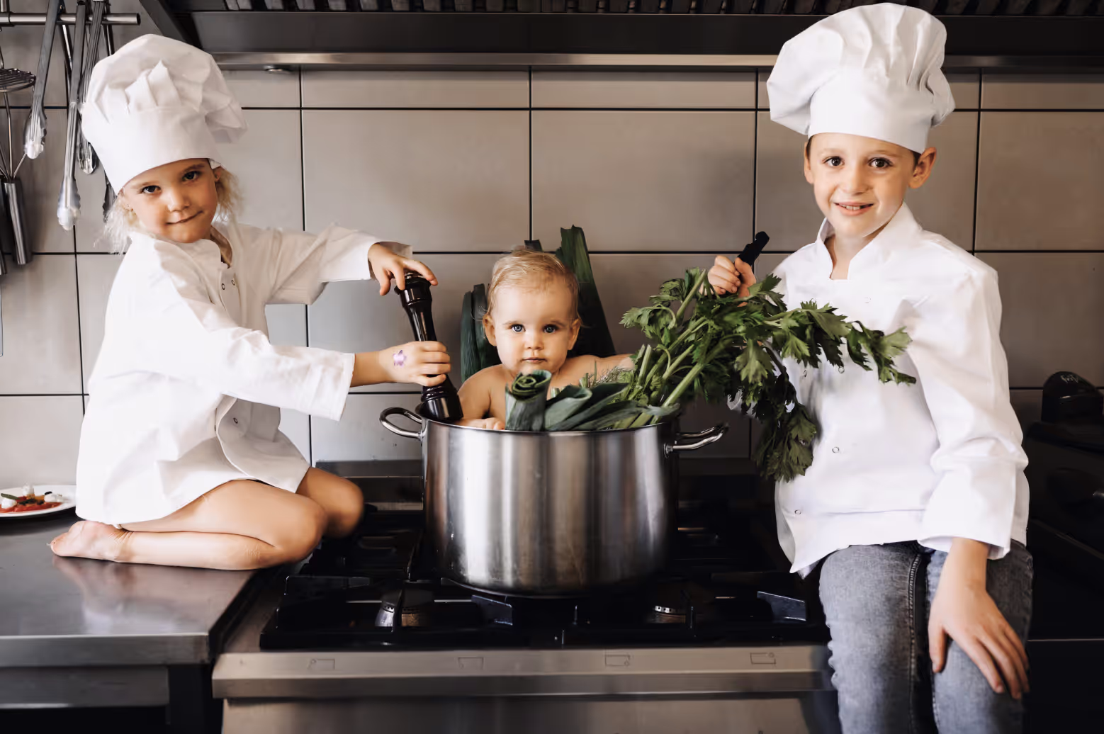 Twee kinderen in koksoutfits met een baby tussen groenten in een grote kookpot op het fornuis.