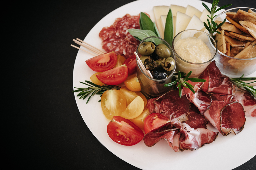 Plateerbord met plakjes vleeswaren, tomaten, kaas, olijven, dip en knapperige crackers.