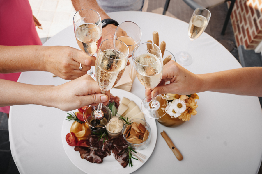 Vijf handen klinken met glazen champagne boven een tafel met een bord hapjes en bloemen.