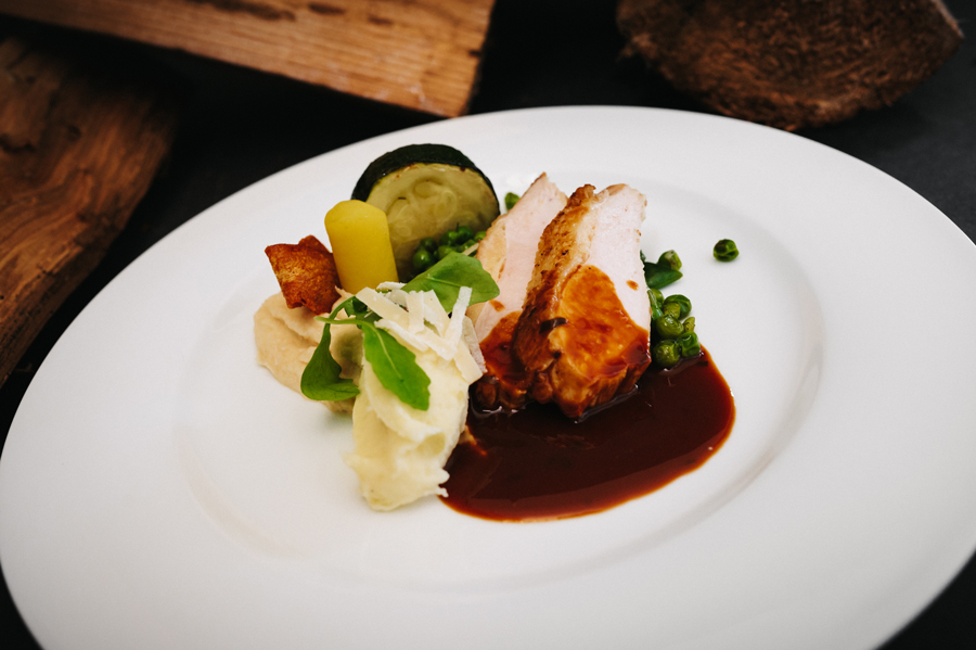 Geroosterde kip met saus, peentjes, doperwten, courgette en puree op een wit bord.
