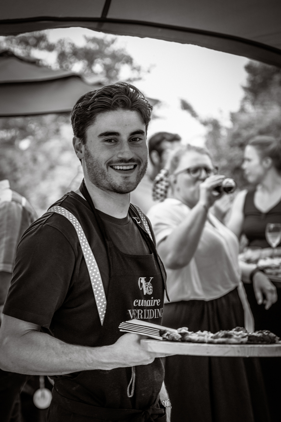 Jonge man met schort die lacht en een dienblad met eten vasthoudt op een outdoor evenement.