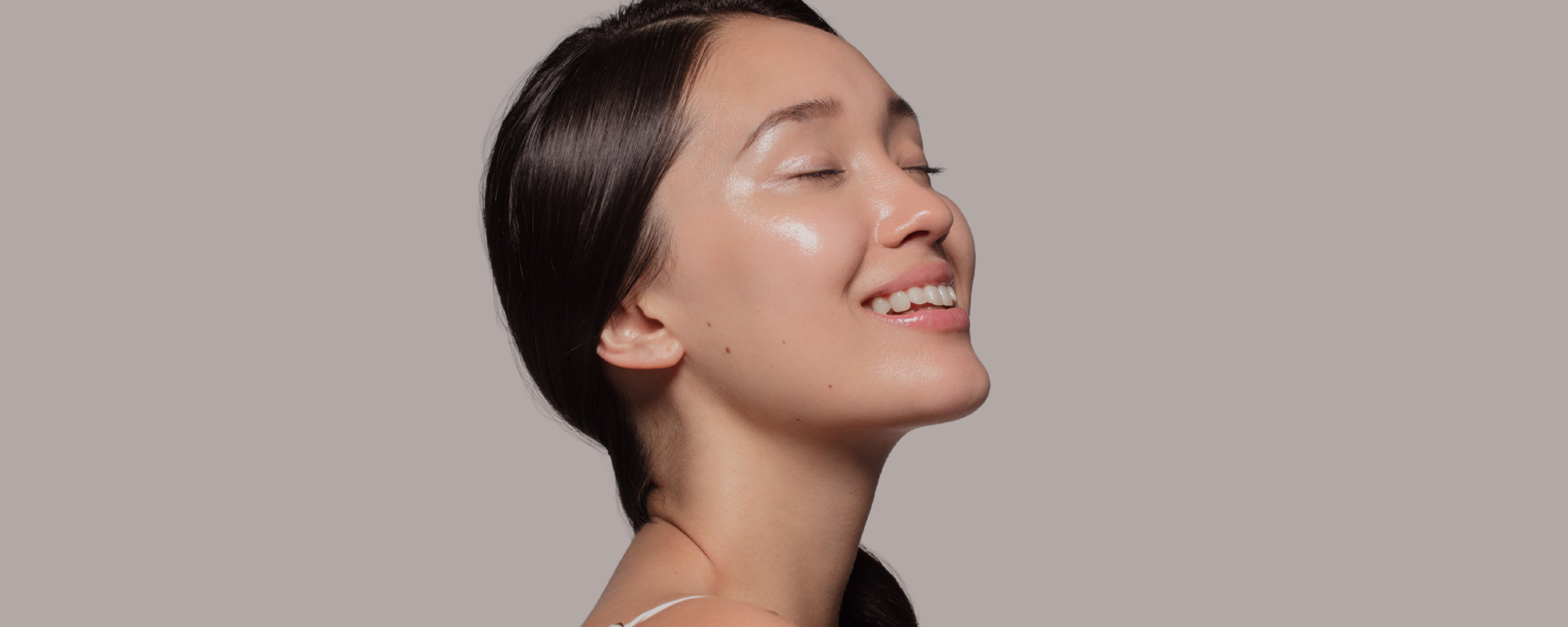 Side profile of a smiling woman with clear, glowing skin and dark hair against a light taupe background.