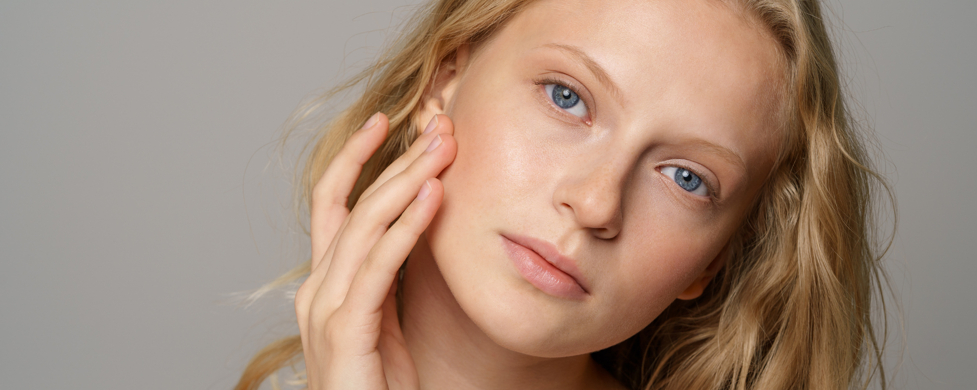 Close-up of a blonde woman with blue eyes touching her smooth skin on her cheek.